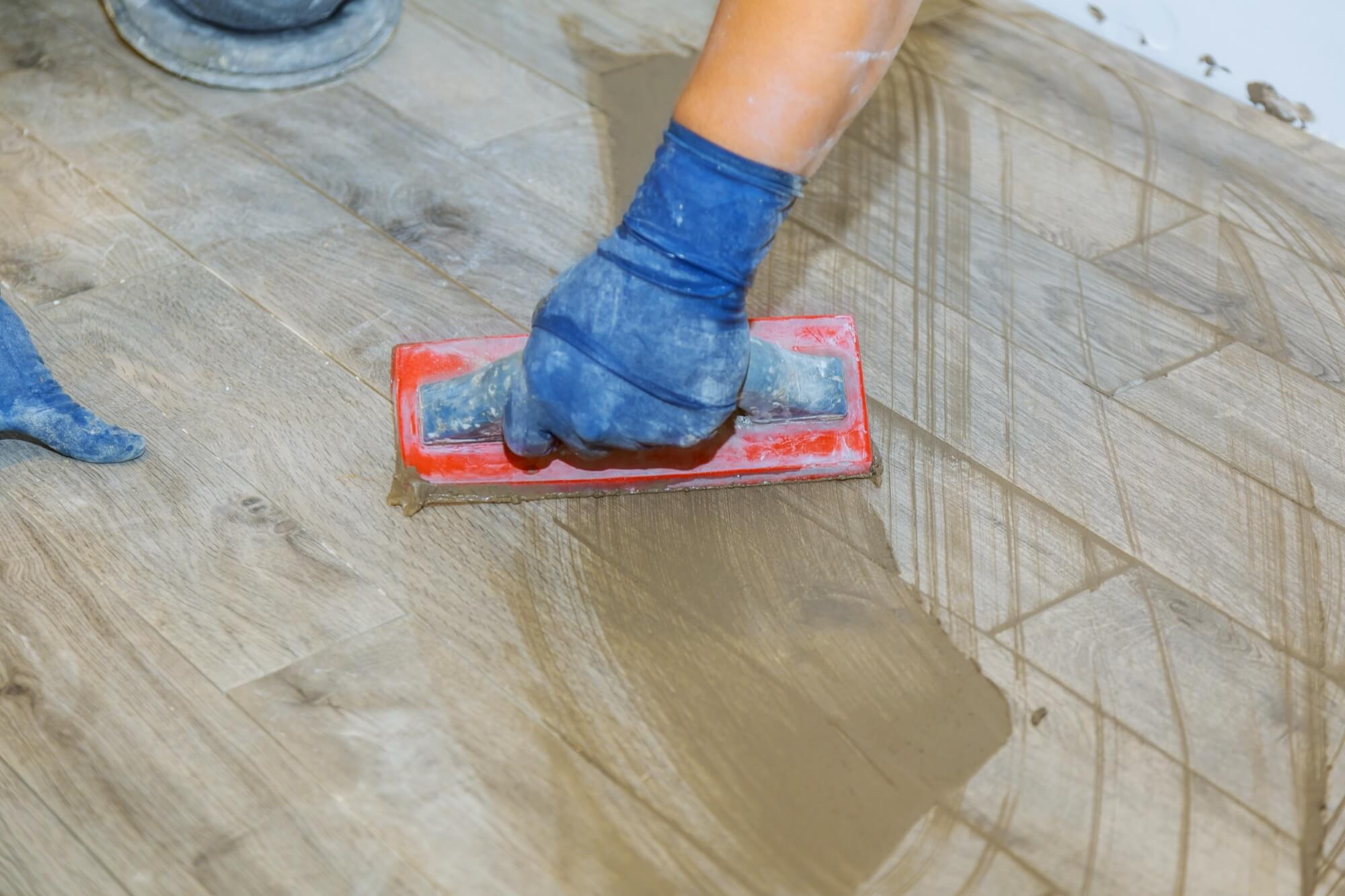 Närbild av en hantverkare som fogar ett trägolv under en renovering, iförd blå handskar.